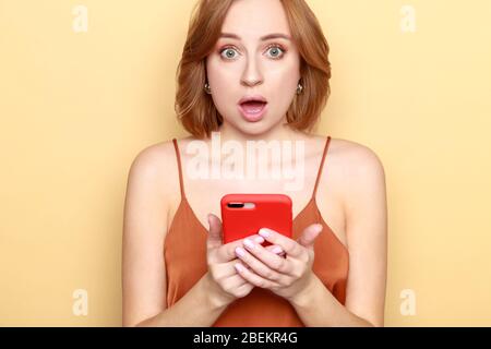 Portrait of shocked or anxious young woman holding smartphone, looking at camera with open mouth, has surprised facial expression, shocked by prices, Stock Photo