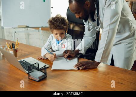 Caucasian teenboy as a doctor consulting for patient, giving recommendation, treating. Little doctor during working with older colleague. Concept of childhood, human emotions, health, medicine. Stock Photo