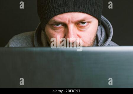 Man wearing black cap and hoodie sitting behind the computer monitor in the dark, close up, hacking computer system concept Stock Photo