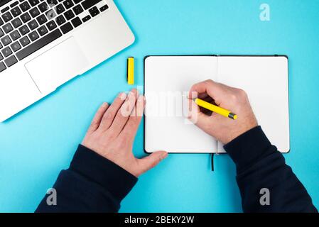 directly above shot of person taking notes with pen and paper on blue desk with laptop computer Stock Photo