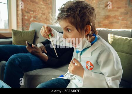 Caucasian teenboy as a doctor consulting for patient at home, giving recommendation, treating. Little doctor taking temperature, shocked. Concept of childhood, human emotions, health, medicine. Stock Photo