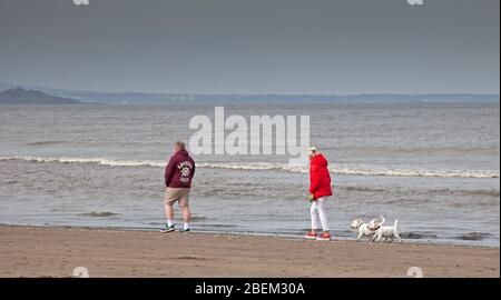 Portobello, Edinburgh, Scotland, UK. 14th April 2020. A hazy cloudy scene at the very quiet seaside due to the Coronavirus Lockdown, with mostly dog walkers. Pictured this couple walking along the shore with two West Highland Terrriers. Stock Photo