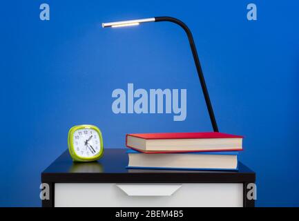 A modern lamp, an alarm clock and two books on a nightstand against a blue colored interior wall Stock Photo