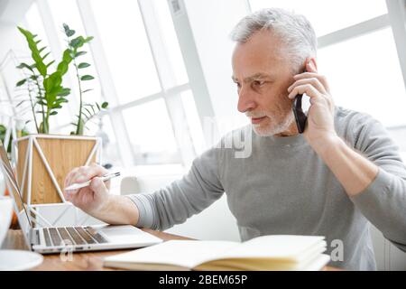Photo of serious mature man making notes while working with laptop and cellphone in cafe indoors Stock Photo