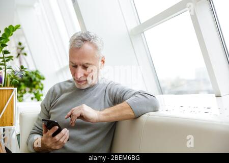 Picture of a handsome mature senior grey-haired bearded businessman sit in cafe using mobile phone. Stock Photo