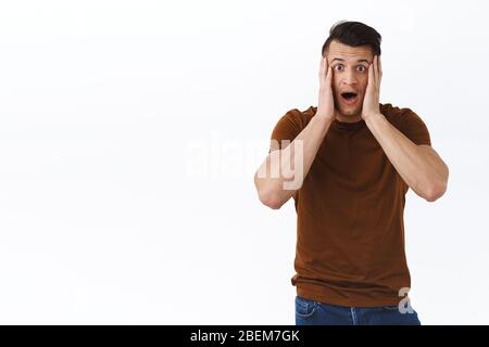 Portrait of shocked and speechless young handsome man, hold hands on face, open mouth and staring camera impressed with disbelief, hear overwhelming Stock Photo