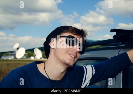 Young Caucasian man with sunglasses leaning on the car roof and smiling Selective focus Stock Photo