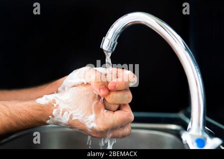 Wash your hands. a young guy washes his hands with soap under the faucet over a metal sink on a black dark background. Virus prevention. Hygienic proc Stock Photo
