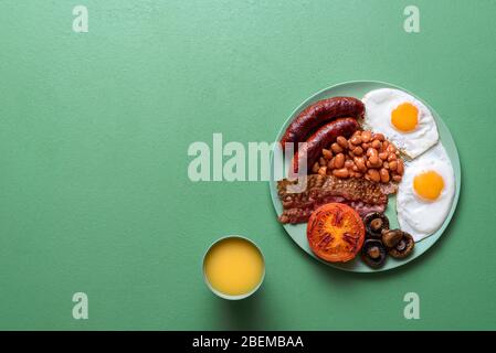 Traditional english breakfast with baked beans, sausages, bacon and sunny-side-up eggs on a green background. British full breakfast on a plate. Stock Photo