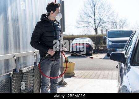 washing car by water pressure at self-service station. A man with a respirator on his face protects himself from a viral epidemic Stock Photo