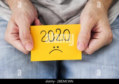 Man holding yellow piece of paper with the year 2020 and a sad smiley on it in his hands.  2020 horrible year concept. Close up. Stock Photo