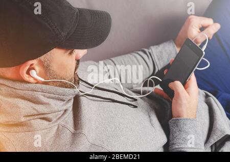 Portrait of handsome bearded man in black cap watching videos or listening to the music with earphones on smartphone. Stock Photo