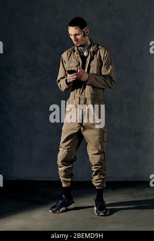 Stylish tattooed man with earrings, beard. Dressed in khaki overalls, black sneakers. Using smartphone, posing on gray background. Dancehall, hip-hop Stock Photo