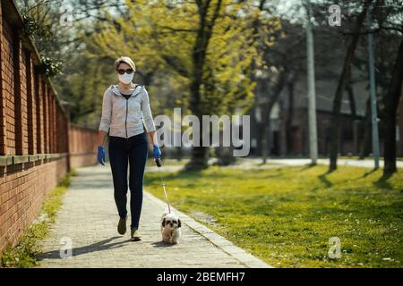A woman in a medical protective mask is walking along the city street path with her dear cute little Shih Tzu dog during flu virus outbreak and corona Stock Photo