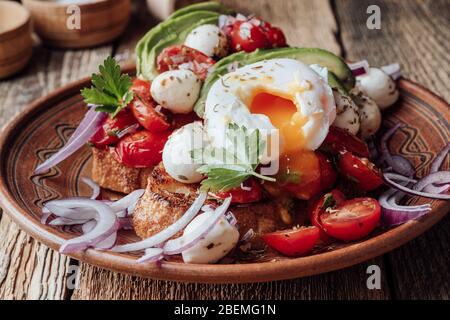 Tomato toast with avocado, mozzarella, poached egg and herbs in rural ceramic bowl, close up Stock Photo