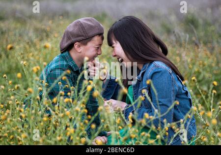 Pregnant Asian mother with long black hair wearing a green dress and blue denim jacket her son in a brown newsboy cap laughing together in a meadow Stock Photo