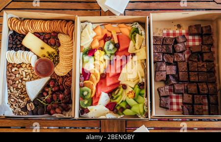 Three boxes with chocolate brownies, tropical fruits and vegetarian antipasto for snacks on wooden table. Stock Photo