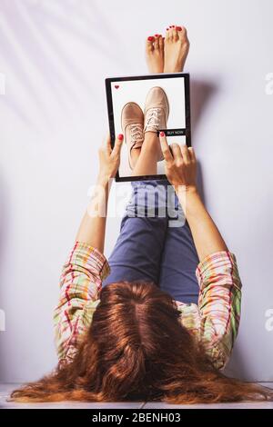 Woman lays with legs up on the wall and pushes buy now button on a laptop buying shoes. Online shopping concept.Self isolation. Stock Photo