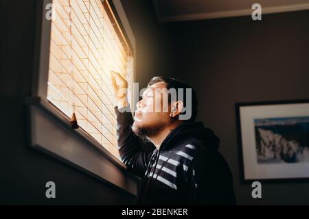 Asian man in isolation looking through window blinds. Man forced to stay inside the house as a result of the restrictions caused by the Coronavirus ou Stock Photo