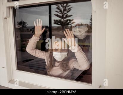 COVID-19 Lockdown. Depressed lonely little girl with face mask looking through the window during quarantine. Sad sick child in self isolation at home. Stock Photo