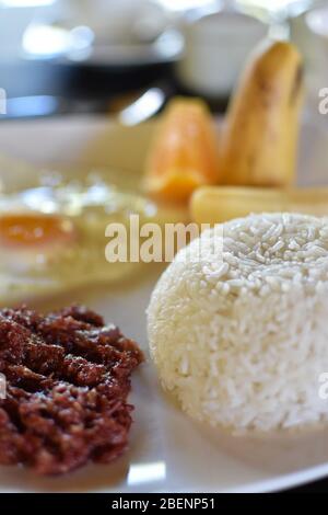 A plate of breakfast consisting of white rice, corned beef, fried egg, and sliced fruits Stock Photo