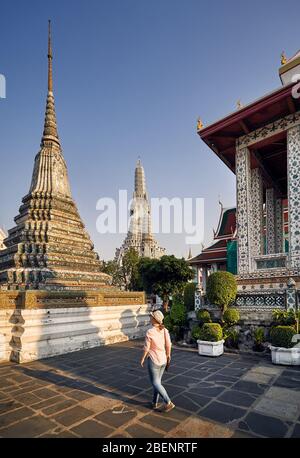 Beautiful tourist woman in hat and pink shirt walking in Wat Arun temple at sunset in Bangkok, Thailand.