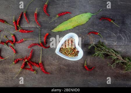 studio shot of assorted spices with whole red peppers rosemary and allium ursinum with melted mixture of them in heart shaped white bowl  on wooden bo