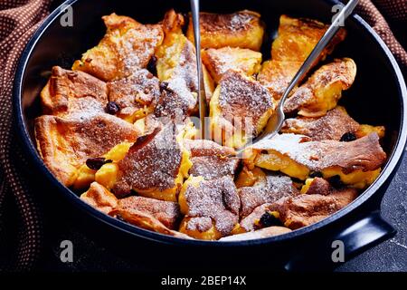 Light and fluffy austrian pancake kaiserschmarrn with raisins and icing sugar on top, cutlery on a black baking dish on a dark concrete table, horizon Stock Photo