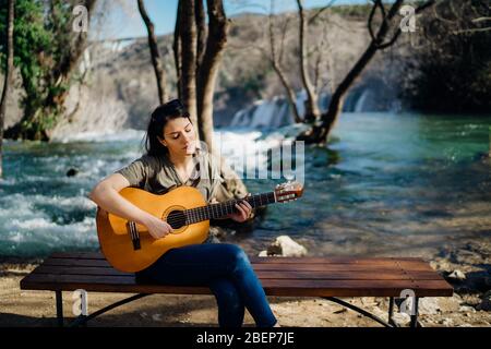Young guitarist playing acoustic guitar and looking to river nature.Searching inspiration.Music creator.New artist in good mood.Musical talent.Smiling Stock Photo