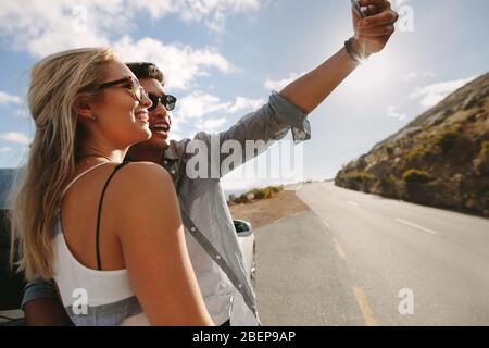 Couple taking a selfie using a smart phone and smiling while standing near car during travel. Man and woman posing for a self portrait by their car wh Stock Photo