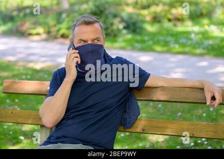 Man wearing a face guard using his mobile phone as he relaxes outdoors on a park bench in spring during the coronavirus or Covid-19 pandemic Stock Photo