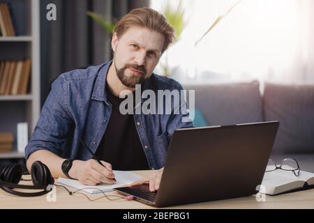 Bearded man in denim shirt sitting at home with opened laptop and looking at camera. Mature person working on distance during isolation. Stock Photo