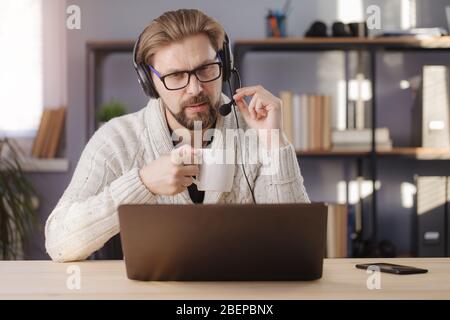 Serious bearded man in eyeglasses and knitted sweater looking at computer screen while sitting at table. Mature male in headset having video call whil Stock Photo