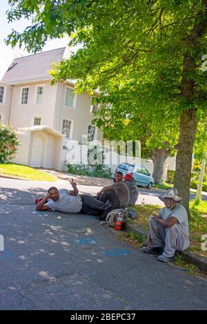 Black building workers rest on the roadside during the day. They are working in an exclusive and upmarket area of Cape Town called Claremont. The men are friendly and happy. There is a nice mansion behind them and a beautiful tree. Both bask in the sunshine. The residents are mostly white and they create a lot of work for the community. Stock Photo