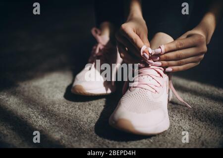 Young woman tying shoelace on sporty sneakers before trainings at home. Concept of active and healthy lifestyle. Close up, dark atmosphere. Stock Photo