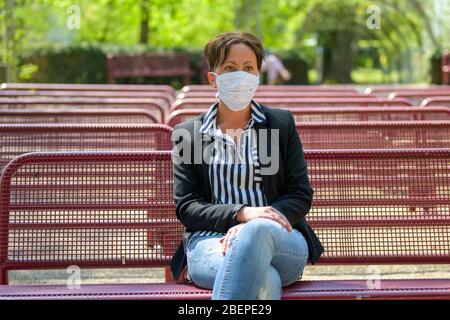 Stylish woman wearing a face mask in a park as she relaxes at an outdoor venue with rows of empty benches during the coronavirus or Covid-19 pandemic Stock Photo