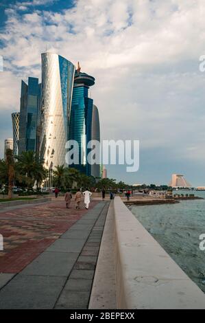 People walk along Al Corniche with the Doha Tower  and modern buildings of West Bay in the background. Stock Photo