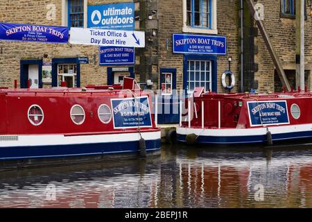 2 bright red blue canal narrow boats moored on water by boat hire business ticket office & banner - Leeds-Liverpool Canal, Skipton, Yorkshire, England Stock Photo