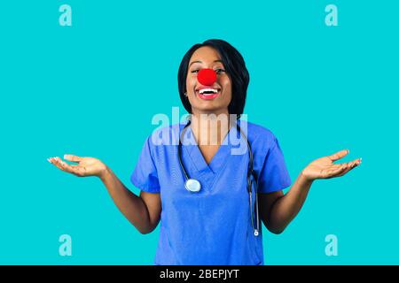 Portrait of a smiling female doctor or nurse wearing blue scrubs uniform and red nose with arms out isolated on blue background Stock Photo