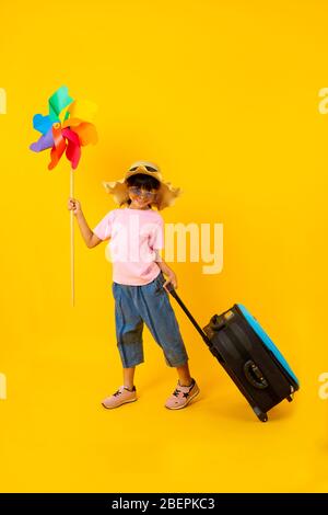 Portrait of young Asian pretty girl holding colorful turbine and drag blue travel bag, Thai kid in summer style on yellow background Stock Photo