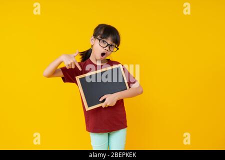 Young Asian girl student in red shirt holding blank small blackboard to present words on yellow background, pretty kid wearing eye glasses pointing an Stock Photo