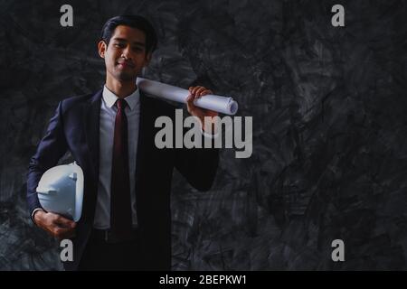 Young business engineering holding white helmet and blue print on dark background, smart and confident worker. Stock Photo