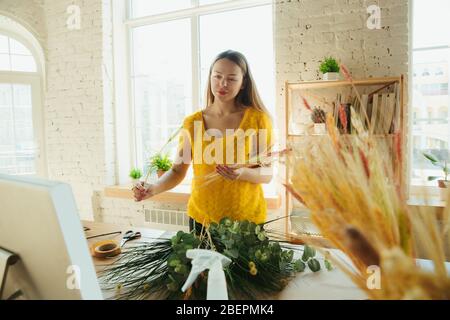 Florist at work: woman shows how to make bouquet. Young caucasian woman gives online workshop of doing gift, present for celebration, with computer. Working at home while isolated, quarantined concept. Stock Photo