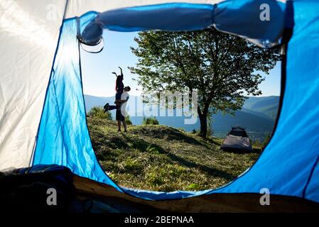Back view of romantic couple tourists resting near camping and big tree in the mountains at sunny morning. Man holding woman in hands. View from inside tent. Tourism adventure active lifestyle concept Stock Photo