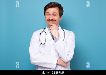 Medical doctor in white coat smiling at camera rubbing his chin posing against blue background Stock Photo