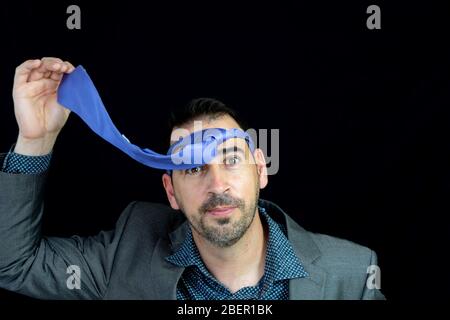 Bearded young man dressed in gray suit posing with tie on head after night party with black background Stock Photo