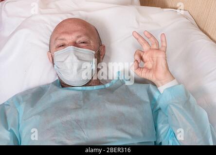 An elderly patient in a protective mask during a pandemic in bed shows approx. Fighting anxiety and caring for health. Social distance during the epid Stock Photo