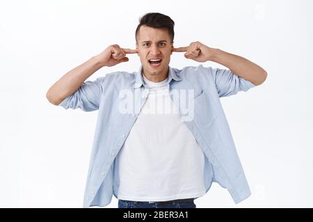 Turn volume off. Portrait of annoyed, bothered handsome man look irritated at camera, shut ears with fingers from annoying bothering sound, loud music Stock Photo