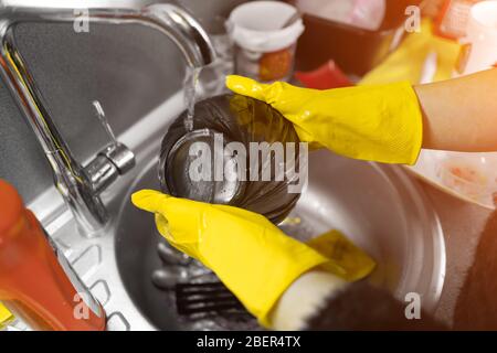 Close up of female hands in yellow protective rubber gloves washing white plate with blue cleaning sponge against kitchen sink full of foam and tablew