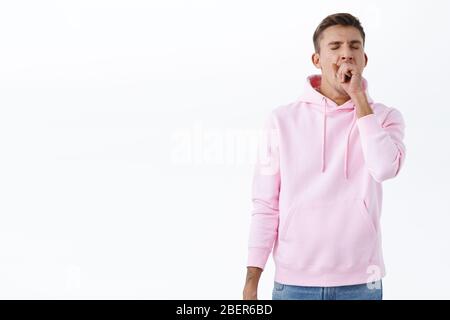 Portrait of sleepy handsome blond man in pink hoodie, close eyes and yawning over fatigue, tired of working late, waking-up morning, cover opened Stock Photo